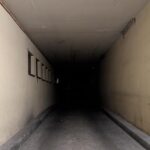 Ein enger, schwach beleuchteter Tunnel mit cremefarbenen Wänden und kleinen vergitterten Fenstern auf der linken Seite, der in die Dunkelheit führt. (KI generierte Beschreibung)