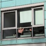 Eine Person sitzt an einem Schreibtisch in einem Gebäude, das durch ein großes Fenster mit türkisfarbenen Außenwänden betrachtet wird. (KI generierte Beschreibung)