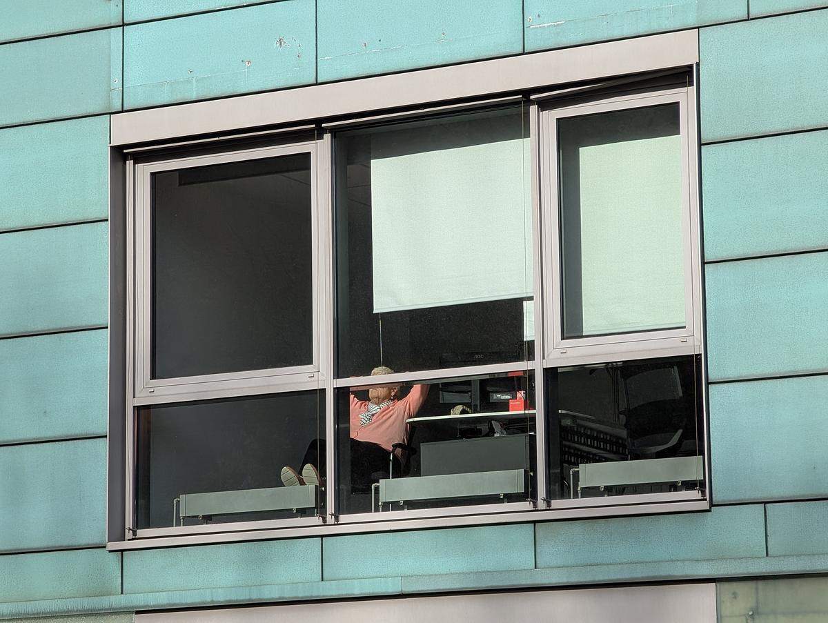 Eine Person sitzt an einem Schreibtisch in einem Gebäude, das durch ein großes Fenster mit türkisfarbenen Außenwänden betrachtet wird. (KI generierte Beschreibung)