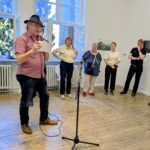 Ein Mann mit Hut liest aus einer Zeitung in ein Mikrofon, während hinter ihm in einem hellen, großen Raum mit großen Fenstern fünf Personen im Halbkreis stehen. (KI generierte Beschreibung)