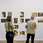 Zwei Personen stehen vor einer weißen Wand, an der mehrere sepiafarbene Fotos in einer Kunstgalerie ausgestellt sind. (KI generierte Beschreibung)