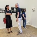 Drei Personen befinden sich in einer Kunstgalerie; zwei stehen und unterhalten sich, während eine Person die Fotos an der Wand betrachtet. (KI generierte Beschreibung)