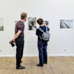 Zwei Personen stehen vor einer weißen Wand und betrachten und besprechen ausgestellte Schwarz-Weiß-Fotografien; eine hält eine Kamera, während die andere auf ein Bild zeigt. (KI generierte Beschreibung)