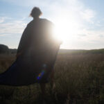 Eine Person steht auf einer Wiese, ihr Gewand weht im Wind; die tief stehende Sonne strahlt von hinten und lässt sie als Silhouette erscheinen. (KI generierte Beschreibung)