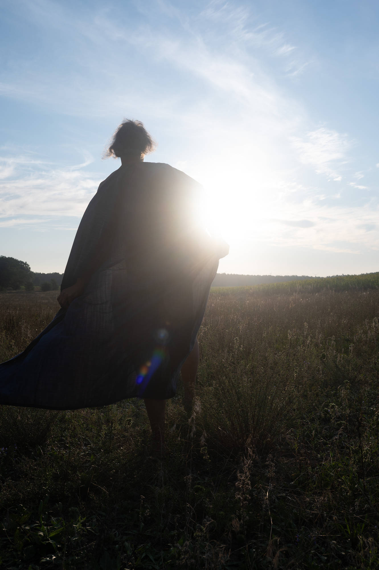 Eine Person steht auf einer Wiese, ihr Gewand weht im Wind; die tief stehende Sonne strahlt von hinten und lässt sie als Silhouette erscheinen. (KI generierte Beschreibung)