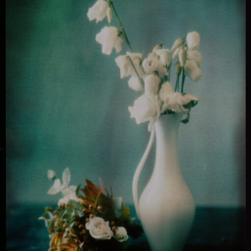 Eine weiße Keramikvase enthält verwelkte weiße Blumen, daneben liegt ein kleiner Strauß verschiedener Blumen auf einer dunklen Fläche vor einem sanften blau-grünen Hintergrund. (KI generierte Beschreibung)