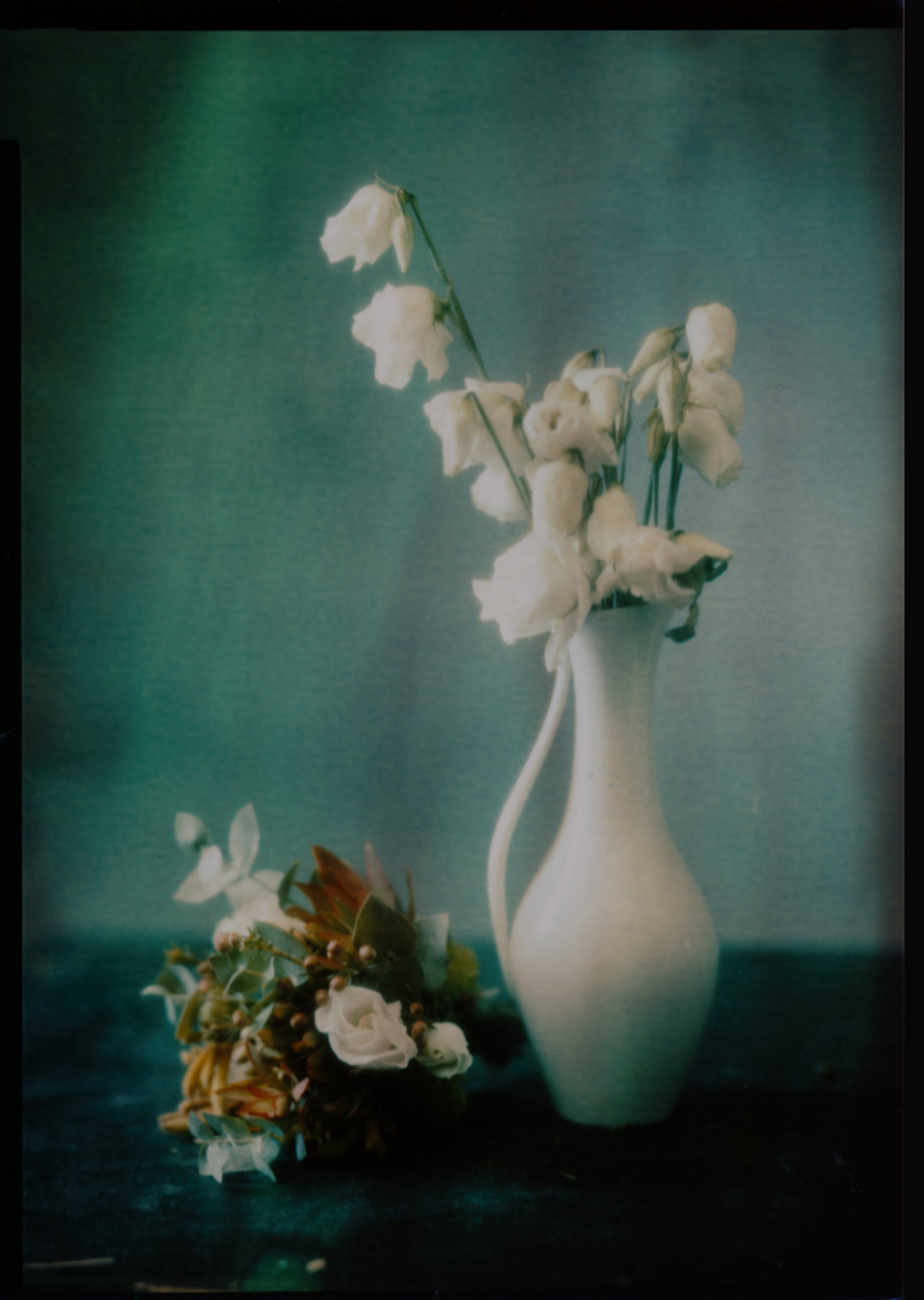 Eine weiße Keramikvase enthält verwelkte weiße Blumen, daneben liegt ein kleiner Strauß verschiedener Blumen auf einer dunklen Fläche vor einem sanften blau-grünen Hintergrund. (KI generierte Beschreibung)