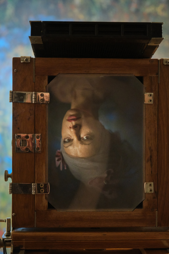 Das Gesicht einer Frau, beleuchtet und auf dem Kopf stehend, spiegelt sich auf der Glasschliffscheibe einer großen Holzfachkamera. (KI generierte Beschreibung)