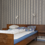 Ein noch nicht bezogenes Holzbett mit blauer Matratze, weißer Bettwäsche und gemusterten Kissen vor einer blumig gestreiften Tapete, darüber zwei Wandleuchten und gerahmte Kunstwerke. (KI generierte Beschreibung)