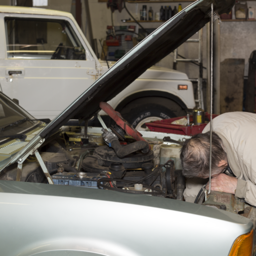 Eine Person in einer beigen Jacke beugt sich in einer Garage über den Motor eines Autos mit geöffneter Motorhaube, während im Hintergrund ein weiteres weißes Fahrzeug geparkt ist. (KI generierte Beschreibung)