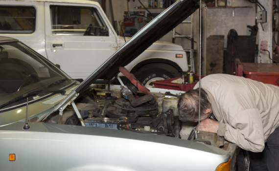 Eine Person in einer beigen Jacke beugt sich in einer Garage über den Motor eines Autos mit geöffneter Motorhaube, während im Hintergrund ein weiteres weißes Fahrzeug geparkt ist. (KI generierte Beschreibung)