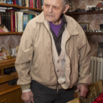 Ein älterer Mann in einer beigen Jacke steht neben einem Esstisch mit roter Tischdecke in einem Raum, der mit Bücherregalen und Dekoartikeln gesäumt ist. (KI generierte Beschreibung)