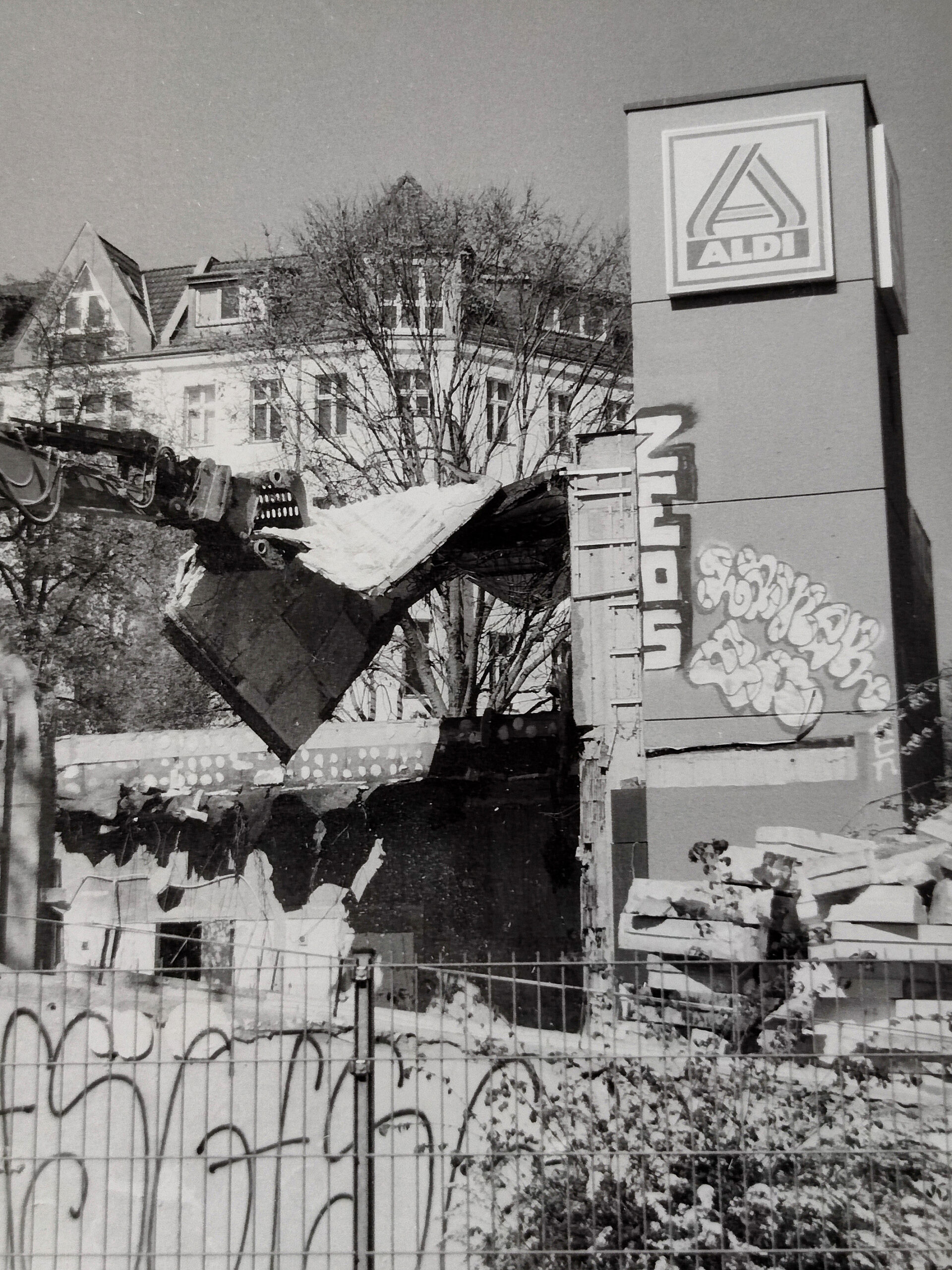 Ein Gebäude mit einem Aldi-Schild wird gerade mit schweren Maschinen abgerissen; an der Seitenwand sind Graffiti zu sehen, und im Hintergrund sind Wohnhäuser zu erkennen. (KI generierte Beschreibung)