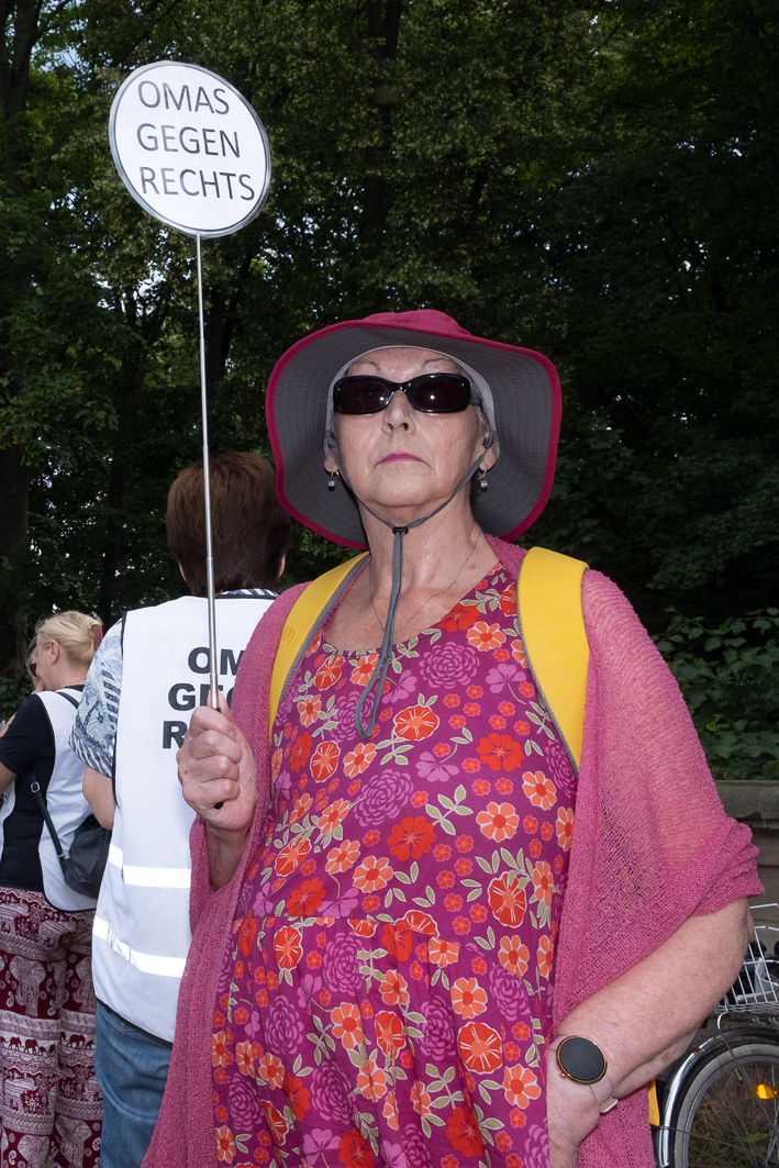 Eine ältere Frau in geblümtem Kleid, Sonnenbrille und Sonnenhut hält bei einer Demonstration ein Schild mit der Aufschrift „OMAS GEGEN RECHTS“. Im Hintergrund sind weitere Teilnehmer und Bäume zu sehen. (KI generierte Beschreibung)