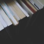 Eine Nahaufnahme der Rücken gestapelter Bücher, aus denen ein Lesezeichen hervorschaut. Alle Bücher stehen auf einem dunklen Regal. (KI generierte Beschreibung)