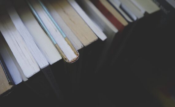 Eine Nahaufnahme der Rücken gestapelter Bücher, aus denen ein Lesezeichen hervorschaut. Alle Bücher stehen auf einem dunklen Regal. (KI generierte Beschreibung)
