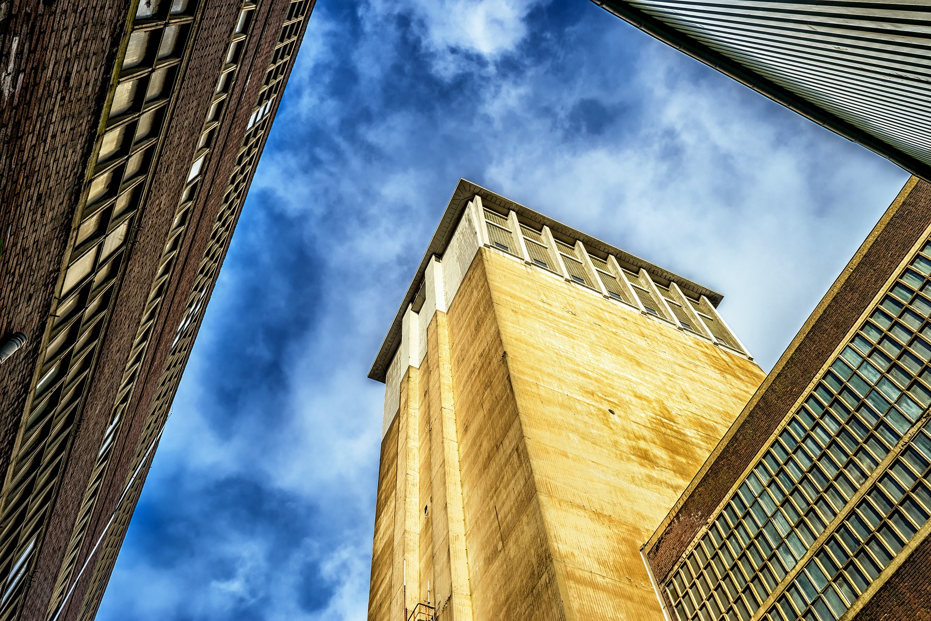 Blick nach oben auf die Ecke eines hoch aufragenden Betongebäudes, das von zwei weiteren hohen Gebäuden unter einem blauen Himmel mit Wolken eingerahmt wird. (KI generierte Beschreibung)