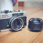Eine Ricoh 500 GX-Kamera steht neben einem separaten Objektiv auf einer Holzoberfläche. (KI generierte Beschreibung)