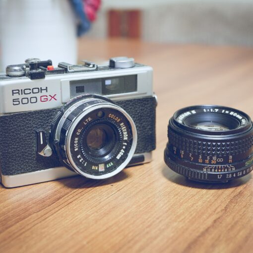 Eine Ricoh 500 GX-Kamera steht neben einem separaten Objektiv auf einer Holzoberfläche. (KI generierte Beschreibung)