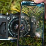 Eine Hand hält ein Samsung-Smartphone und macht damit eine Nahaufnahme von einer alten Zenit-Filmkamera, die im Gras liegt. (KI generierte Beschreibung)