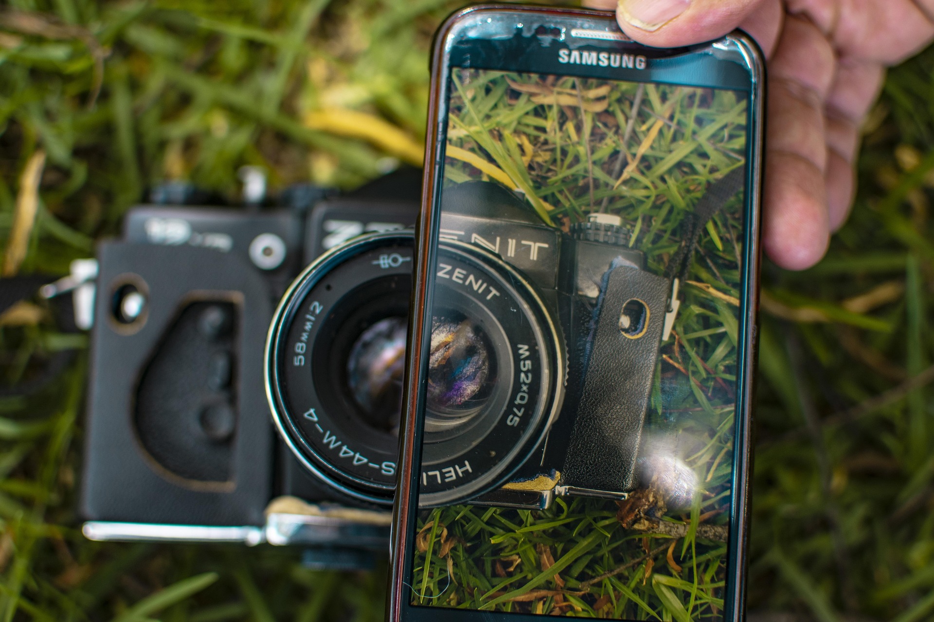 Eine Hand hält ein Samsung-Smartphone und macht damit eine Nahaufnahme von einer alten Zenit-Filmkamera, die im Gras liegt. (KI generierte Beschreibung)