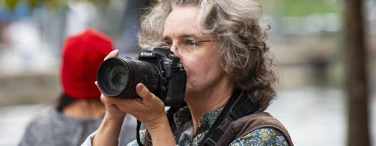 Eine Person mit grauem, lockigem Haar und Brille hält eine Nikon-Kamera an ihr Gesicht und macht ein Foto im Freien. (KI generierte Beschreibung)