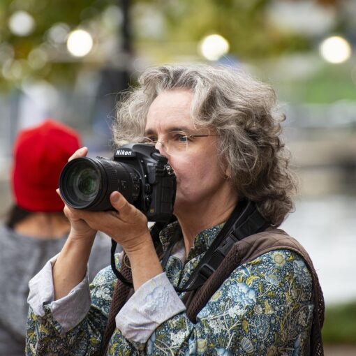 Eine Person mit grauem, lockigem Haar und Brille hält eine Nikon-Kamera an ihr Gesicht und macht ein Foto im Freien. (KI generierte Beschreibung)