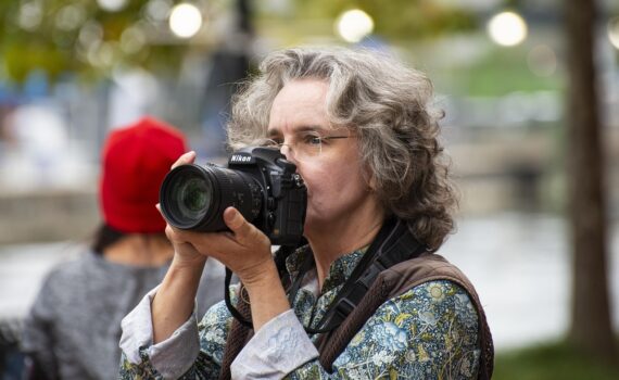 Eine Person mit grauem, lockigem Haar und Brille hält eine Nikon-Kamera an ihr Gesicht und macht ein Foto im Freien. (KI generierte Beschreibung)