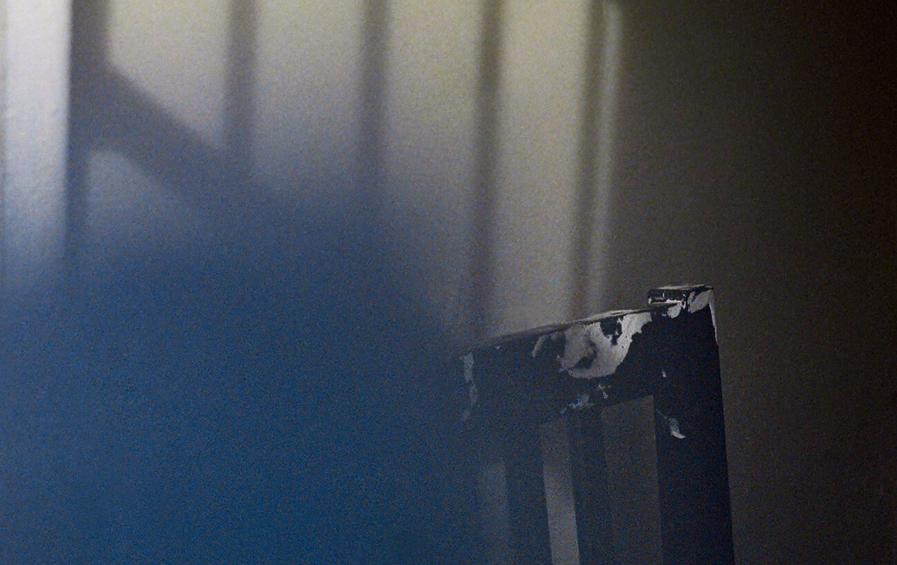 Der Schatten eines Fensterrahmens wird auf eine Wand geworfen. Im schwachen Licht ist ein Stuhl mit abgebrochener Rückenlehne teilweise sichtbar. (KI generierte Beschreibung)