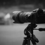 Eine Digitalkamera mit einem großen Teleobjektiv auf einem Stativ wird im Freien aufgestellt und auf einen unscharfen Hintergrund gerichtet. (KI generierte Beschreibung)