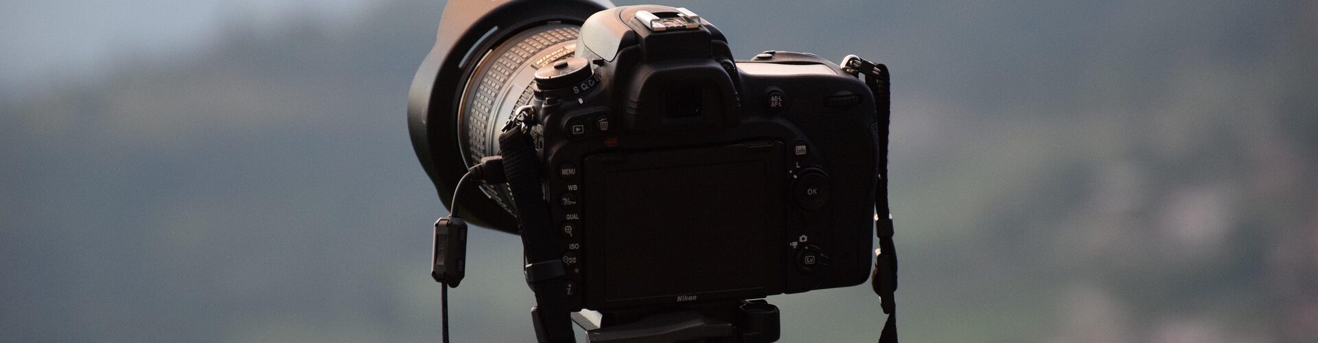 Eine schwarze DSLR-Kamera, die auf einem Stativ montiert ist, steht im Freien und blickt auf eine unscharfe, malerische Landschaft mit Hügeln im Hintergrund. (KI generierte Beschreibung)