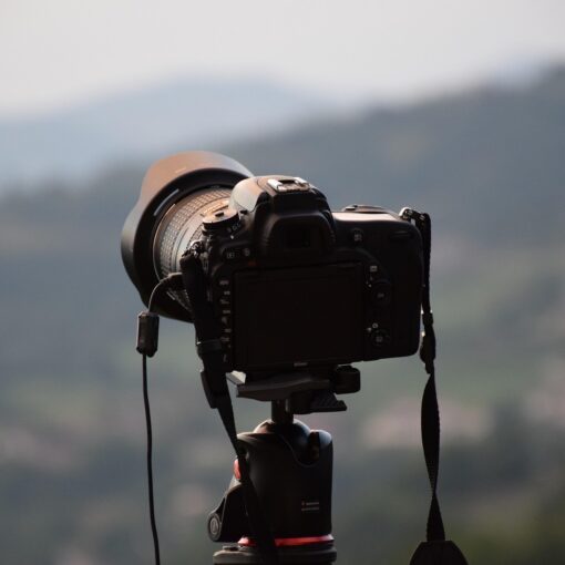 Eine schwarze DSLR-Kamera, die auf einem Stativ montiert ist, steht im Freien und blickt auf eine unscharfe, malerische Landschaft mit Hügeln im Hintergrund. (KI generierte Beschreibung)