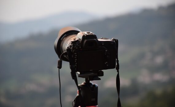 Eine schwarze DSLR-Kamera, die auf einem Stativ montiert ist, steht im Freien und blickt auf eine unscharfe, malerische Landschaft mit Hügeln im Hintergrund. (KI generierte Beschreibung)