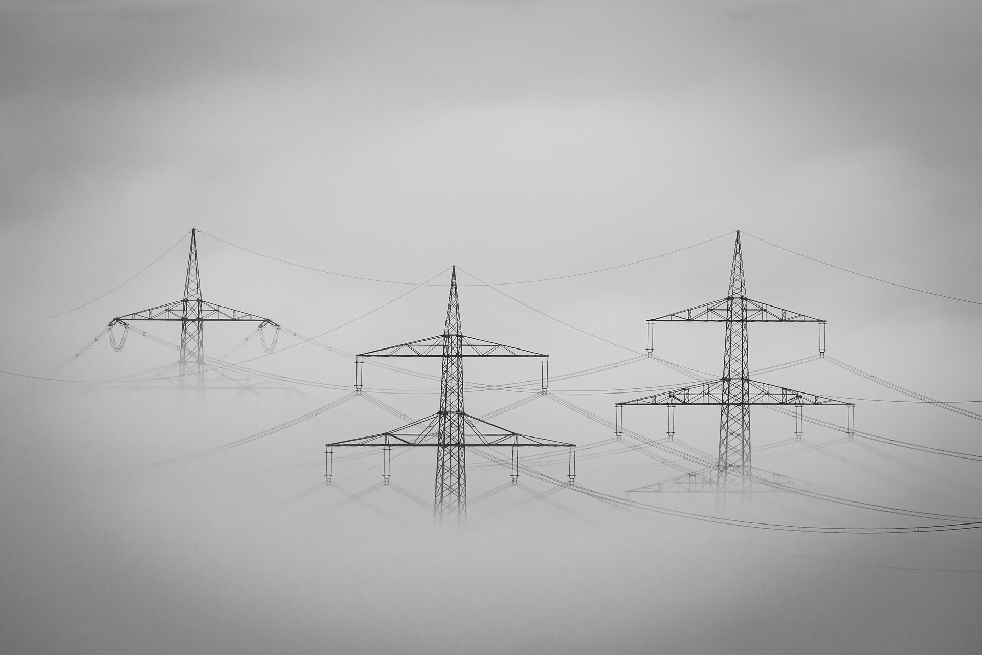 Drei Strommasten tauchen aus dem Nebel auf. Die sichtbaren, miteinander verbundenen Stromleitungen bilden vor einem gedämpften, dunstigen Hintergrund ein symmetrisches Muster. (KI generierte Beschreibung)