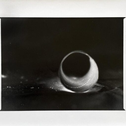 Schwarzweißfoto eines einzelnen Wassertropfens auf einer Oberfläche mit einem dunklen Schatten dahinter. Die Bildränder sind unregelmäßig und weisen auf der rechten Seite Randmarkierungen auf. (KI generierte Beschreibung)