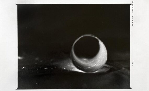 Schwarzweißfoto eines einzelnen Wassertropfens auf einer Oberfläche mit einem dunklen Schatten dahinter. Die Bildränder sind unregelmäßig und weisen auf der rechten Seite Randmarkierungen auf. (KI generierte Beschreibung)