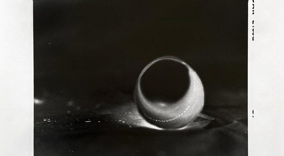Schwarzweißfoto eines einzelnen Wassertropfens auf einer Oberfläche mit einem dunklen Schatten dahinter. Die Bildränder sind unregelmäßig und weisen auf der rechten Seite Randmarkierungen auf. (KI generierte Beschreibung)