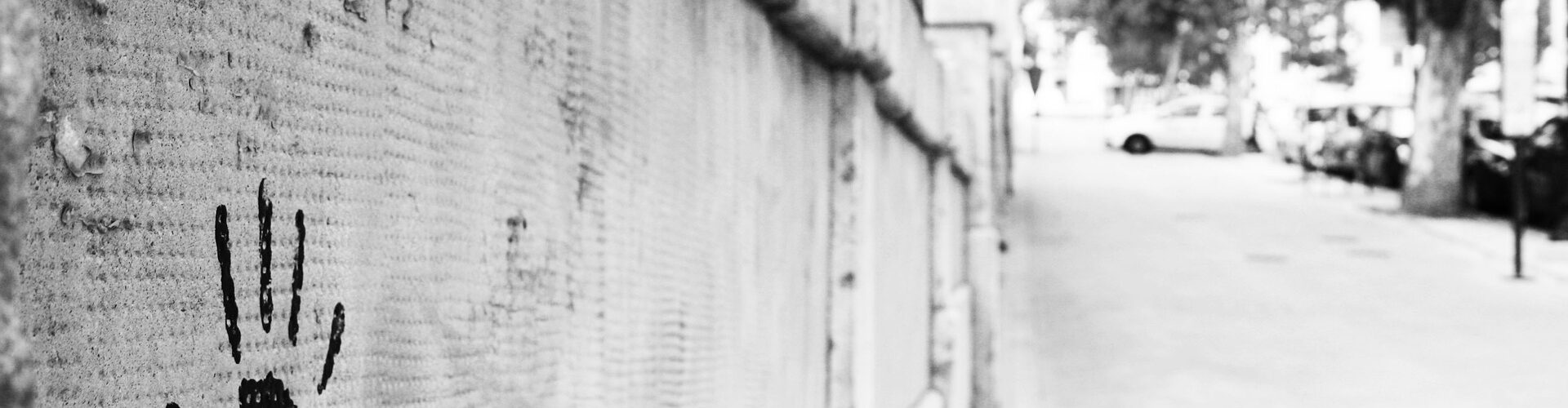 Ein schwarzer Handabdruck und ein Farbspritzer auf einer Betonwand neben einem verlassenen Gehweg, der von Bäumen und geparkten Autos gesäumt ist. (KI generierte Beschreibung)