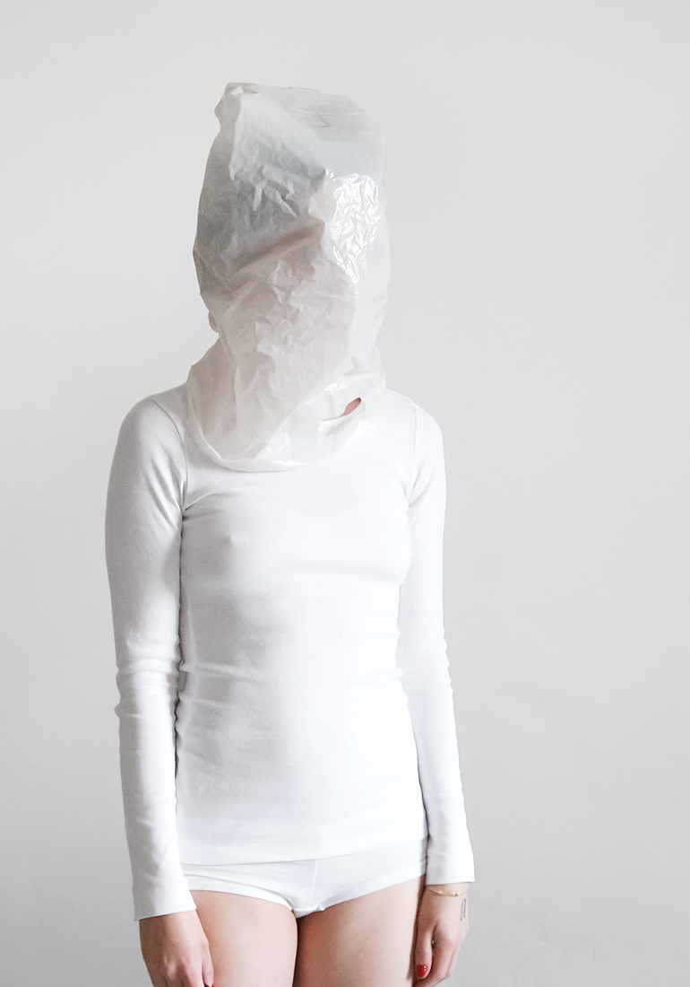 Person in weißem Outfit mit einer durchsichtigen Plastiktüte über dem Kopf, vor einem schlichten weißen Hintergrund stehend. (KI generierte Beschreibung)