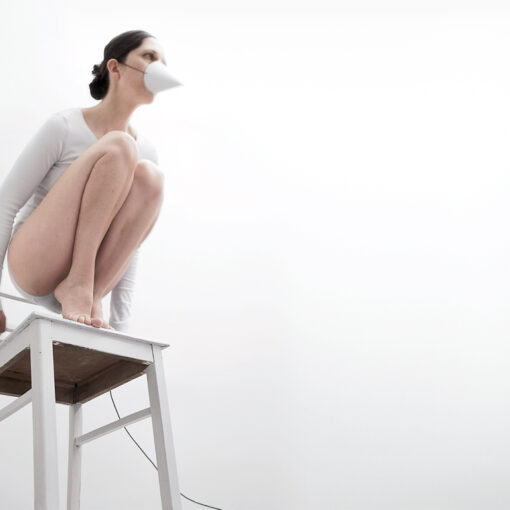 Auf einem weißen Stuhl kauernde Person, die ein weißes Outfit und eine Kegelmaske trägt, vor minimalistischem weißen Hintergrund. (KI generierte Beschreibung)