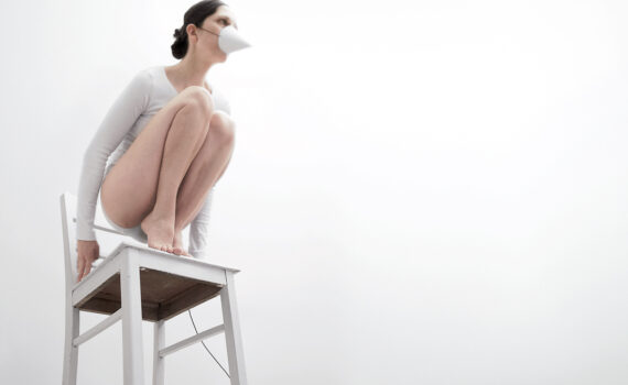 Auf einem weißen Stuhl kauernde Person, die ein weißes Outfit und eine Kegelmaske trägt, vor minimalistischem weißen Hintergrund. (KI generierte Beschreibung)