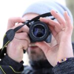 Eine Person mit grauer Beanie und schwarzer Jacke hält eine Kamera vors Gesicht und macht ein Foto. Der Hintergrund ist unscharf. (KI generierte Beschreibung)