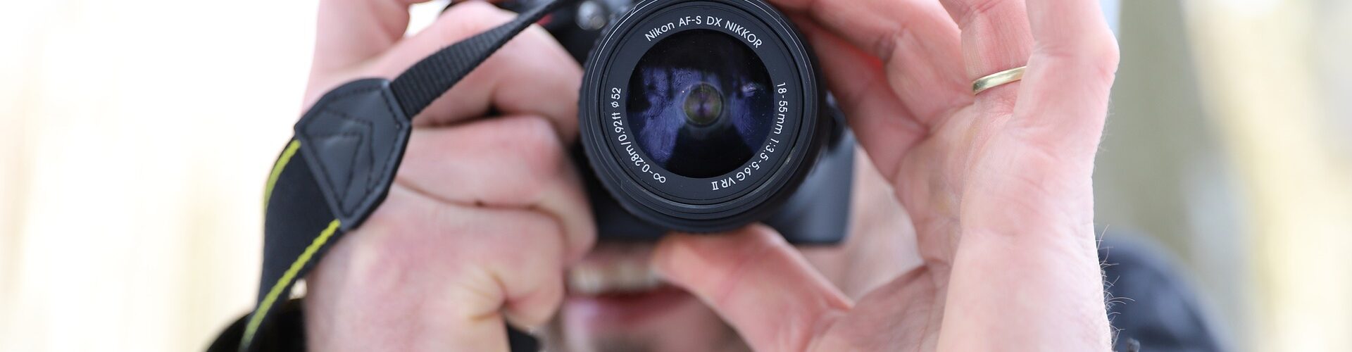 Eine Person mit grauer Beanie und schwarzer Jacke hält eine Kamera vors Gesicht und macht ein Foto. Der Hintergrund ist unscharf. (KI generierte Beschreibung)