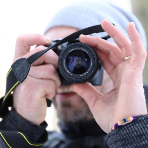 Eine Person mit grauer Beanie und schwarzer Jacke hält eine Kamera vors Gesicht und macht ein Foto. Der Hintergrund ist unscharf. (KI generierte Beschreibung)