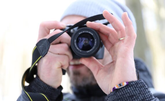 Eine Person mit grauer Beanie und schwarzer Jacke hält eine Kamera vors Gesicht und macht ein Foto. Der Hintergrund ist unscharf. (KI generierte Beschreibung)