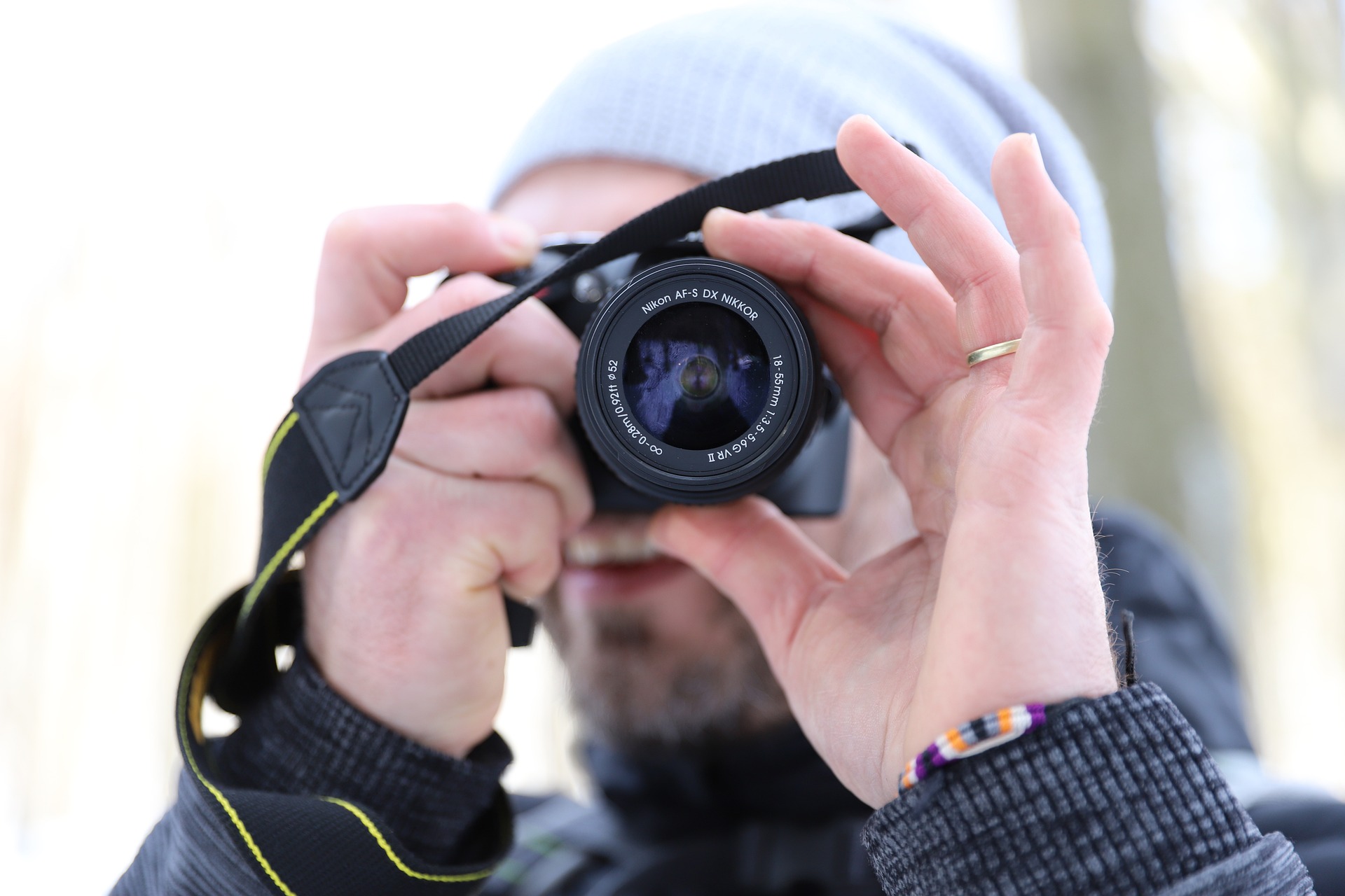 Eine Person mit grauer Beanie und schwarzer Jacke hält eine Kamera vors Gesicht und macht ein Foto. Der Hintergrund ist unscharf. (KI generierte Beschreibung)