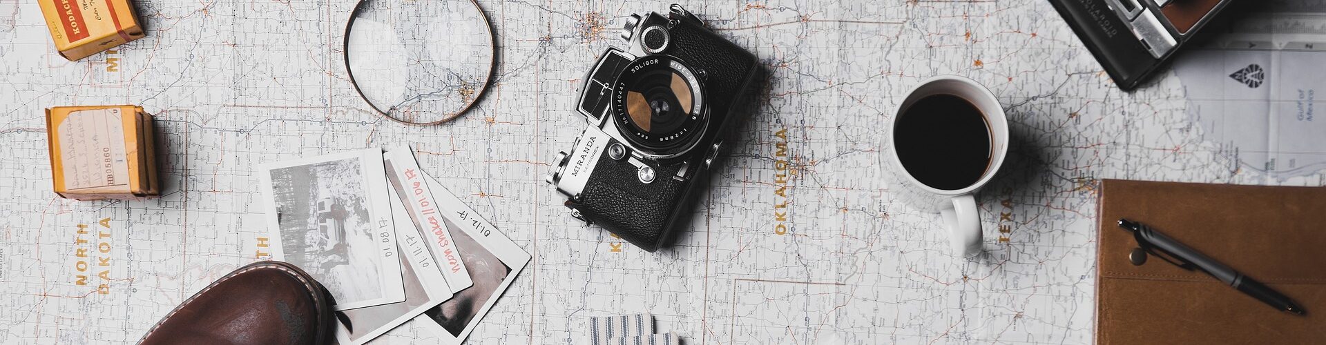 Flache Anordnung der wichtigsten Reiseutensilien, darunter eine Vintage-Kamera, eine Lupe, Filmrollen, eine Karte, eine Kaffeetasse, ein Notizbuch und Stiefel auf einem strukturierten Hintergrund. (KI generierte Beschreibung)