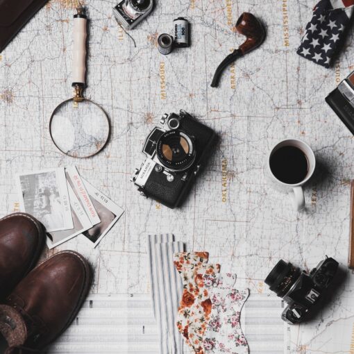 Flache Anordnung der wichtigsten Reiseutensilien, darunter eine Vintage-Kamera, eine Lupe, Filmrollen, eine Karte, eine Kaffeetasse, ein Notizbuch und Stiefel auf einem strukturierten Hintergrund. (KI generierte Beschreibung)