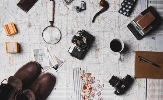 Flache Anordnung der wichtigsten Reiseutensilien, darunter eine Vintage-Kamera, eine Lupe, Filmrollen, eine Karte, eine Kaffeetasse, ein Notizbuch und Stiefel auf einem strukturierten Hintergrund. (KI generierte Beschreibung)