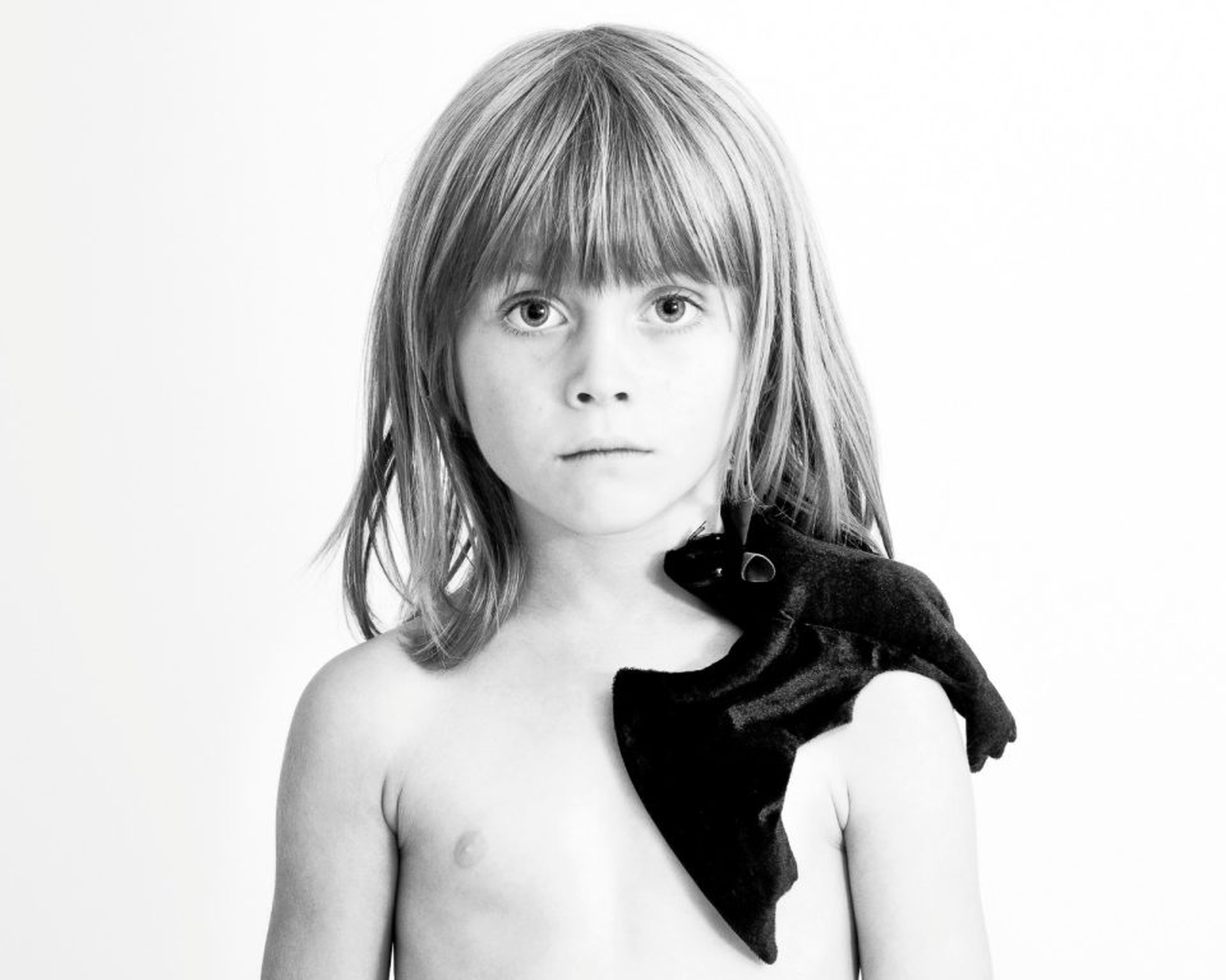Schwarz-Weiß-Foto eines kleinen Kindes mit langen Haaren und nackten Schultern, das ein dunkles Plüschtier über die linke Schulter gelegt hat und vor einem weißen Hintergrund direkt in die Kamera blickt. (KI generierte Beschreibung)